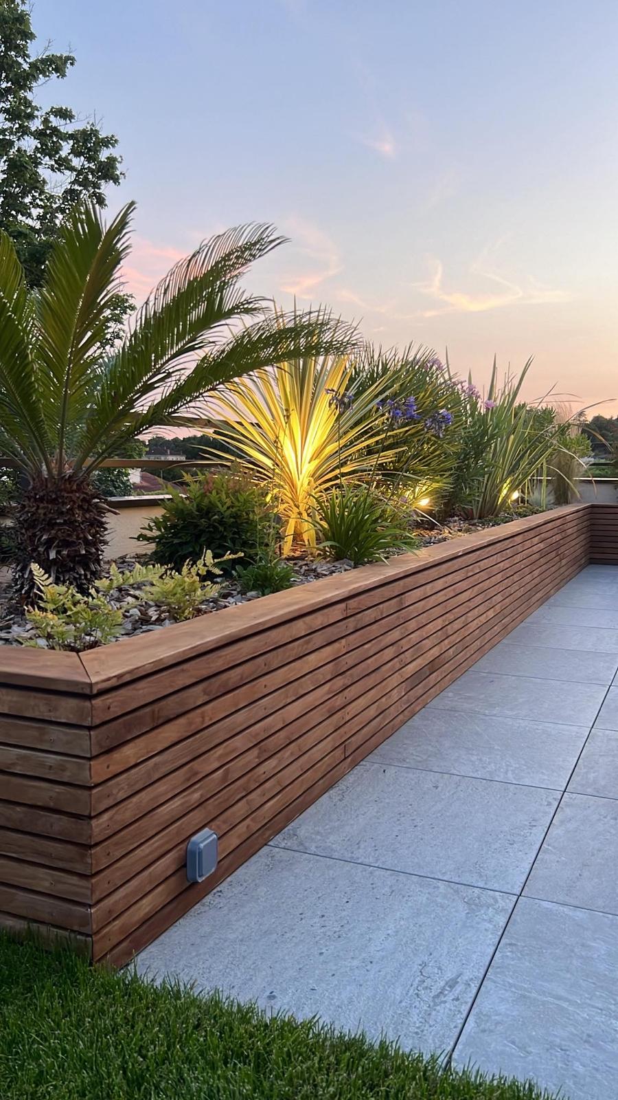 Jardinière en bois sur une terrasse au coucher du soleil, éclairant des plantes exotiques comme un palmier et un yucca.