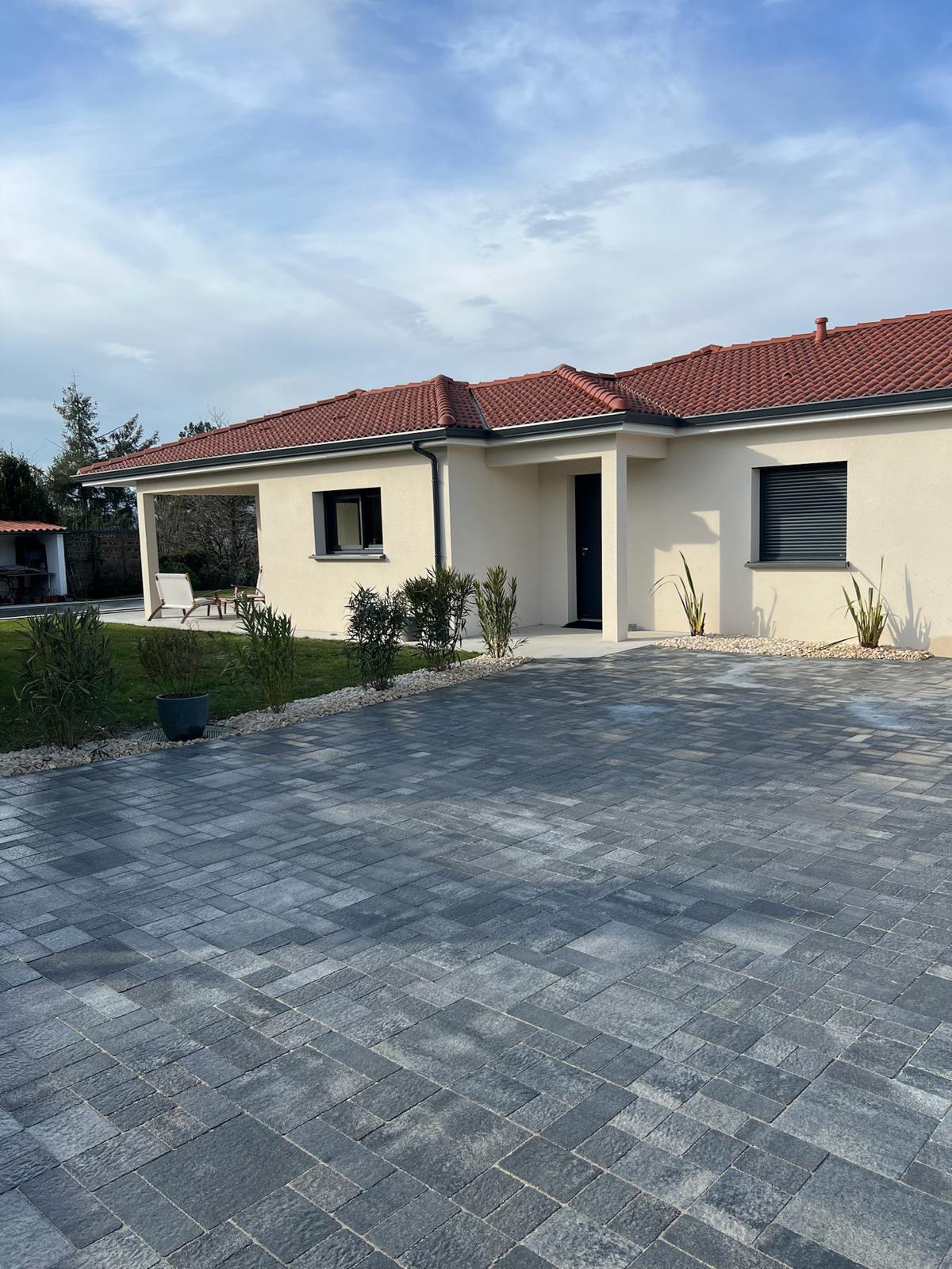 Maison contemporaine de plain-pied avec toiture tuiles rouges, façade crème, porte noire, et grande allée en pavés gris foncé.
