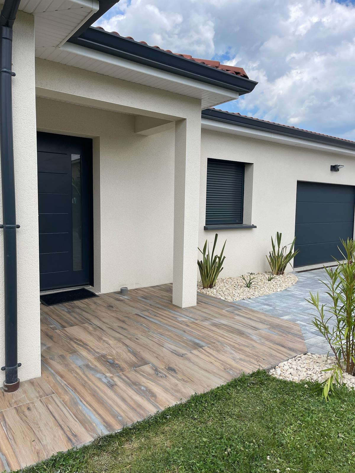 Façade d'une maison moderne avec terrasse en carrelage imitation bois, porte d'entrée anthracite et garage, sous un ciel nuageux.