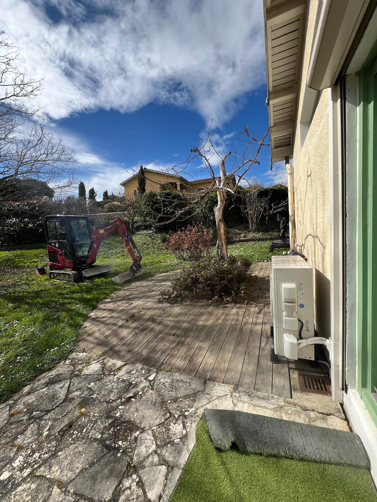 Mini-pelle rouge sur pelouse à côté d'une terrasse en bois, près d'une unité extérieure de climatisation, sous un ciel bleu nuageux.