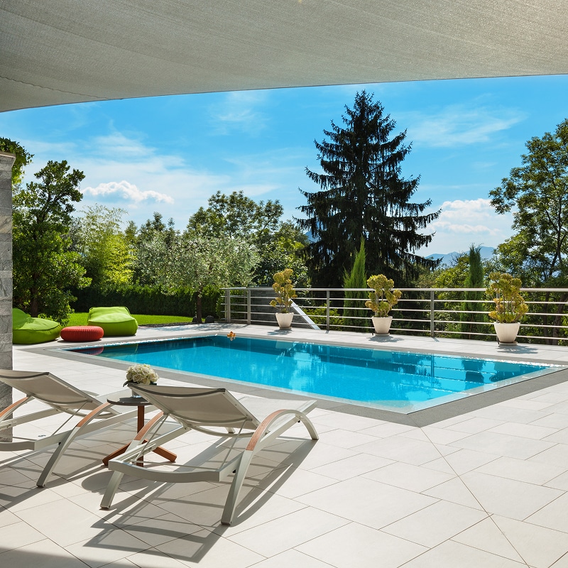 Piscine extérieure moderne sur grande terrasse carrelée, avec deux transats, jardin luxuriant et vue dégagée sous un ciel ensoleillé.