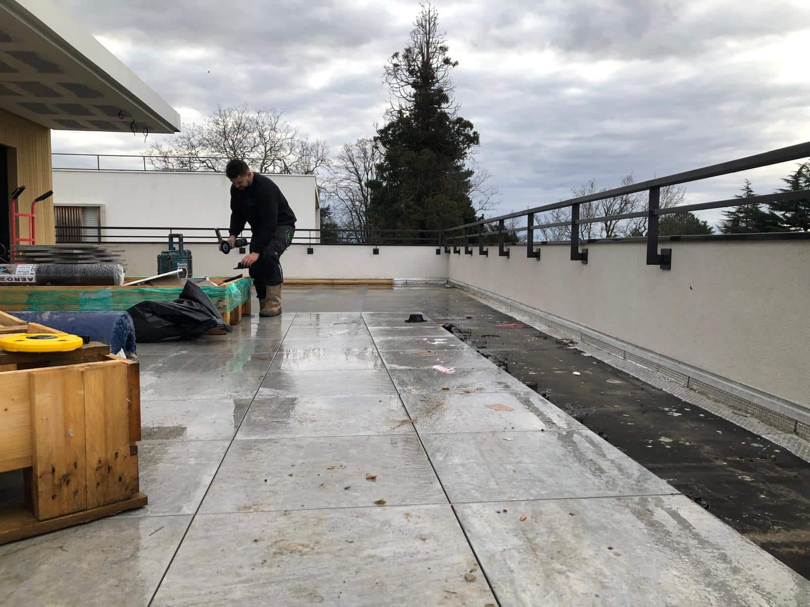 Ouvrier préparant la pose de dalles de terrasse grises sur un toit-terrasse mouillé, avec rambarde noire.