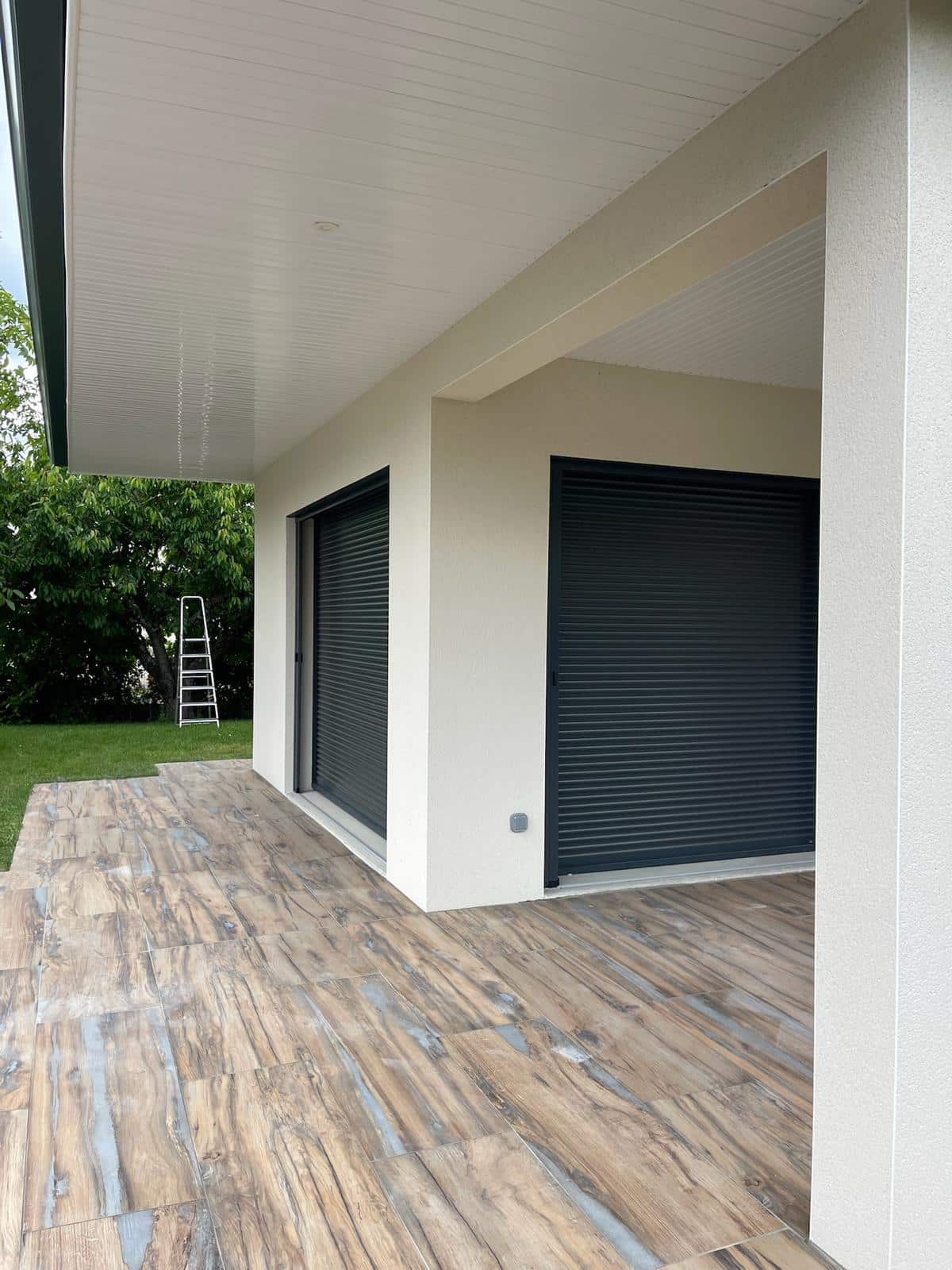 Terrasse moderne avec carrelage imitation bois et volets roulants gris anthracite sur un bâtiment blanc, arrière-plan verdoyant avec échelle.