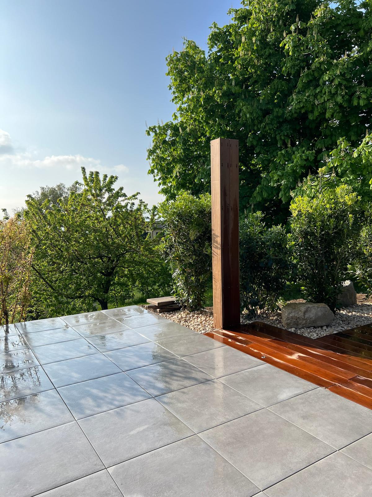 Terrasse moderne avec dalles grises humides et plancher bois brillant, entourée de verdure luxuriante sous un ciel bleu clair.