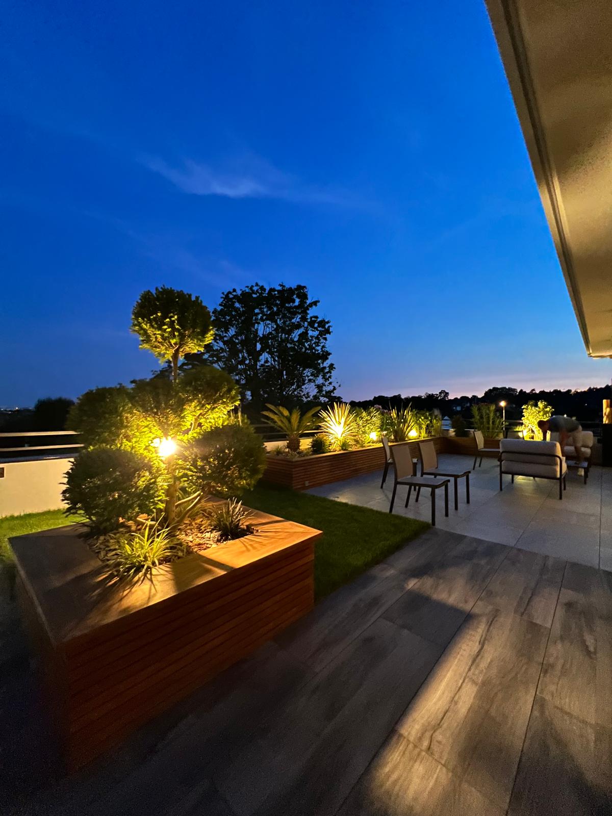 Terrasse moderne au crépuscule avec éclairage chaleureux mettant en valeur des jardinières en bois et des arbres taillés.