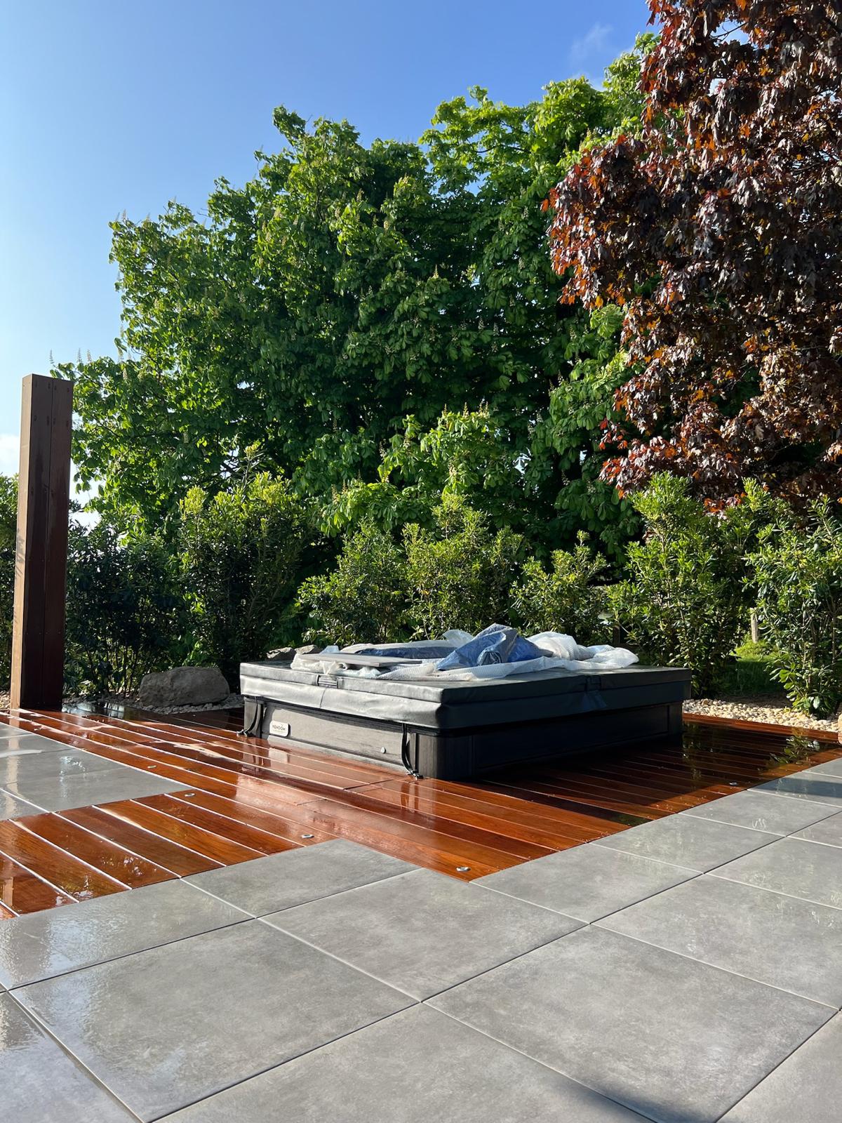 Spa extérieur noir couvert, posé sur une terrasse en bois brillant juxtaposée à des dalles de carrelage grises, entouré d'une végétation luxuriante.