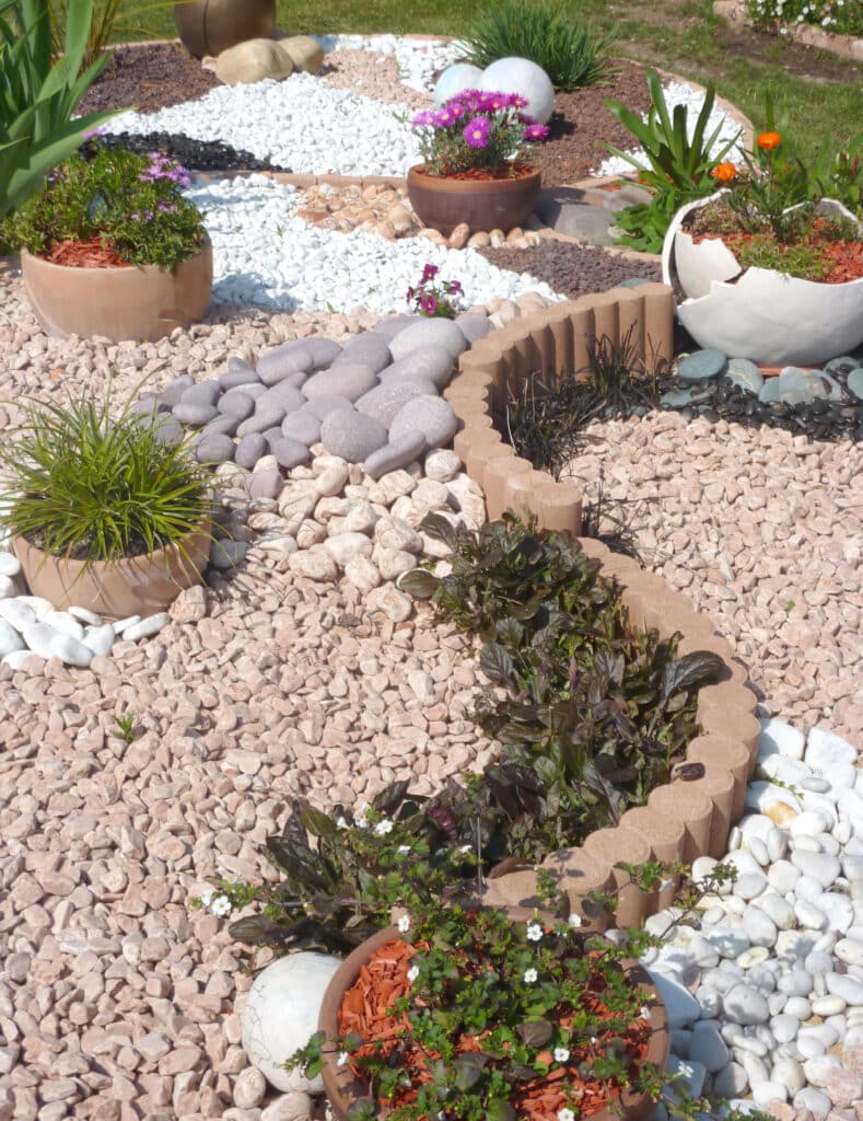 Jardin paysager moderne : graviers décoratifs & fleurs colorées Jardin paysager moderne avec allée de galets gris, graviers blancs et roses, bordures, et plantes en pots fleuris violets et oranges.