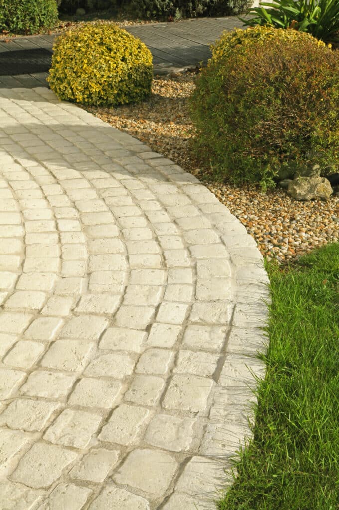 Chemin pavé de jardin: allée courbée & aménagement paysager Chemin pavé clair et courbé dans un jardin, bordé d'herbe verte et de gravier avec des arbustes colorés, menant à une terrasse en bois.