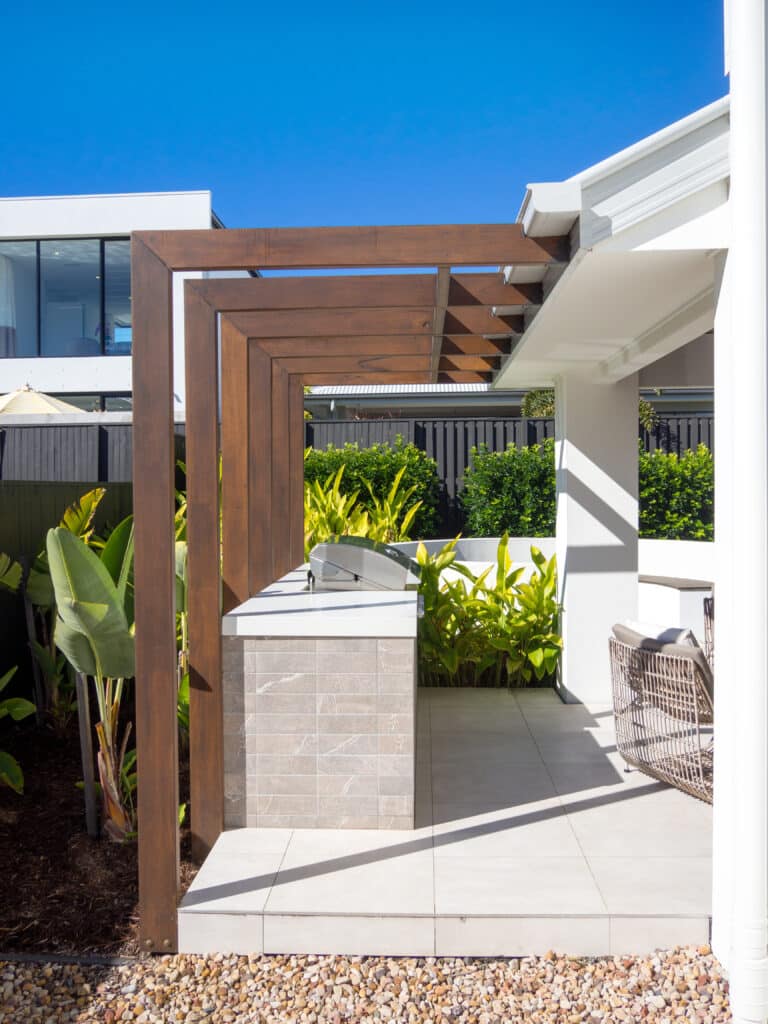 Cuisine extérieure moderne : pergola bois et barbecue intégré Pergola bois et cuisine extérieure moderne avec barbecue intégré, patio carrelé clair, végétation luxuriante et ciel bleu.