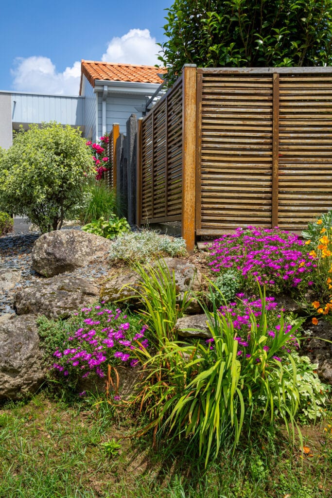 Jardin rocailleux fleuri : aménagement extérieur avec clôture bois. Jardin rocailleux coloré avec fleurs violettes, feuillage vert, grosses pierres, clôture en bois, maison à toit de tuiles sous ciel bleu.