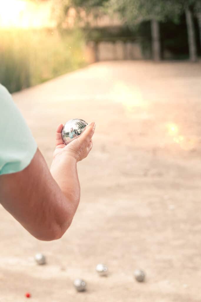 Pétanque : Lancer la boule avec précision en plein air Bras d'un joueur tenant une boule de pétanque brillante, prêt à lancer sur un terrain ensoleillé. D'autres boules et le cochonnet sont au sol.