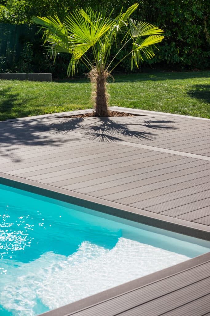 Piscine et terrasse composite: Oasis tropicale avec palmier. Piscine bleue, margelle en bois composite et palmier. Ses feuilles vertes projettent des ombres sur la terrasse et le jardin ensoleillé.