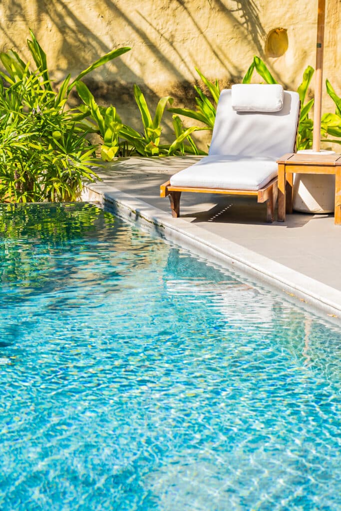 Piscine tropicale et transat : relaxation ensoleillée Piscine bleue scintillante avec transat en bois et coussin blanc, bordée de plantes tropicales luxuriantes et d'un mur ocre au soleil.