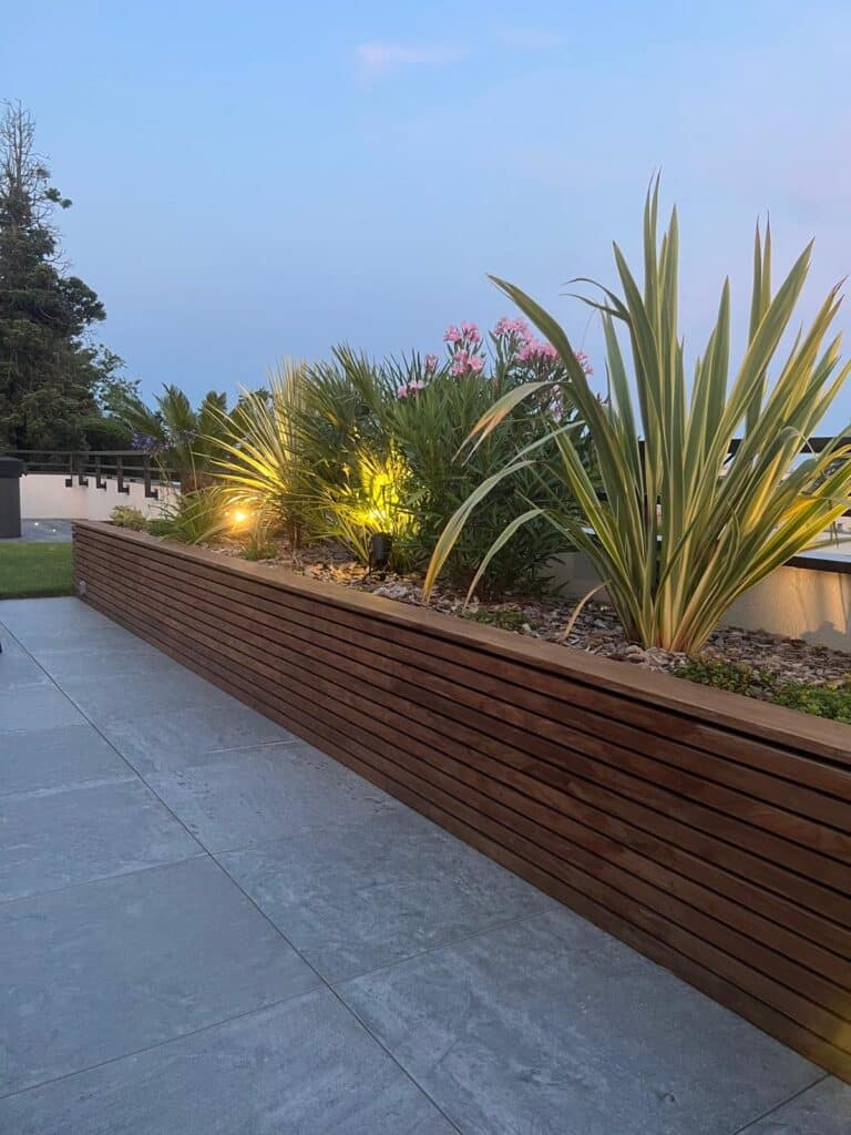 Ambiance Terrasse : Jardinière Bois & Éclairage Végétal Jardinière en bois avec plantes exotiques éclairées par des spots jaunes, au crépuscule, sur une terrasse carrelée grise.
