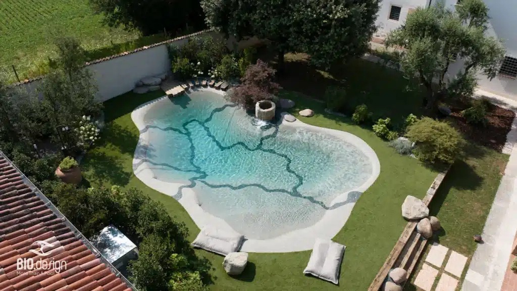 Piscine lagon BIO.design – Créer un jardin naturel. Piscine lagon BIO.design eau turquoise dans un jardin verdoyant. Contour de plage clair, fontaine en pierre et transats doux.