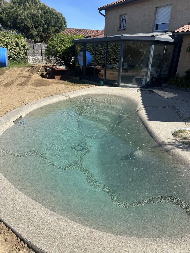 Piscine Lagon Moderne et Véranda : Aménagement Extérieur Piscine lagon avec motifs en galets devant une véranda moderne et un jardin en cours d'aménagement paysager.