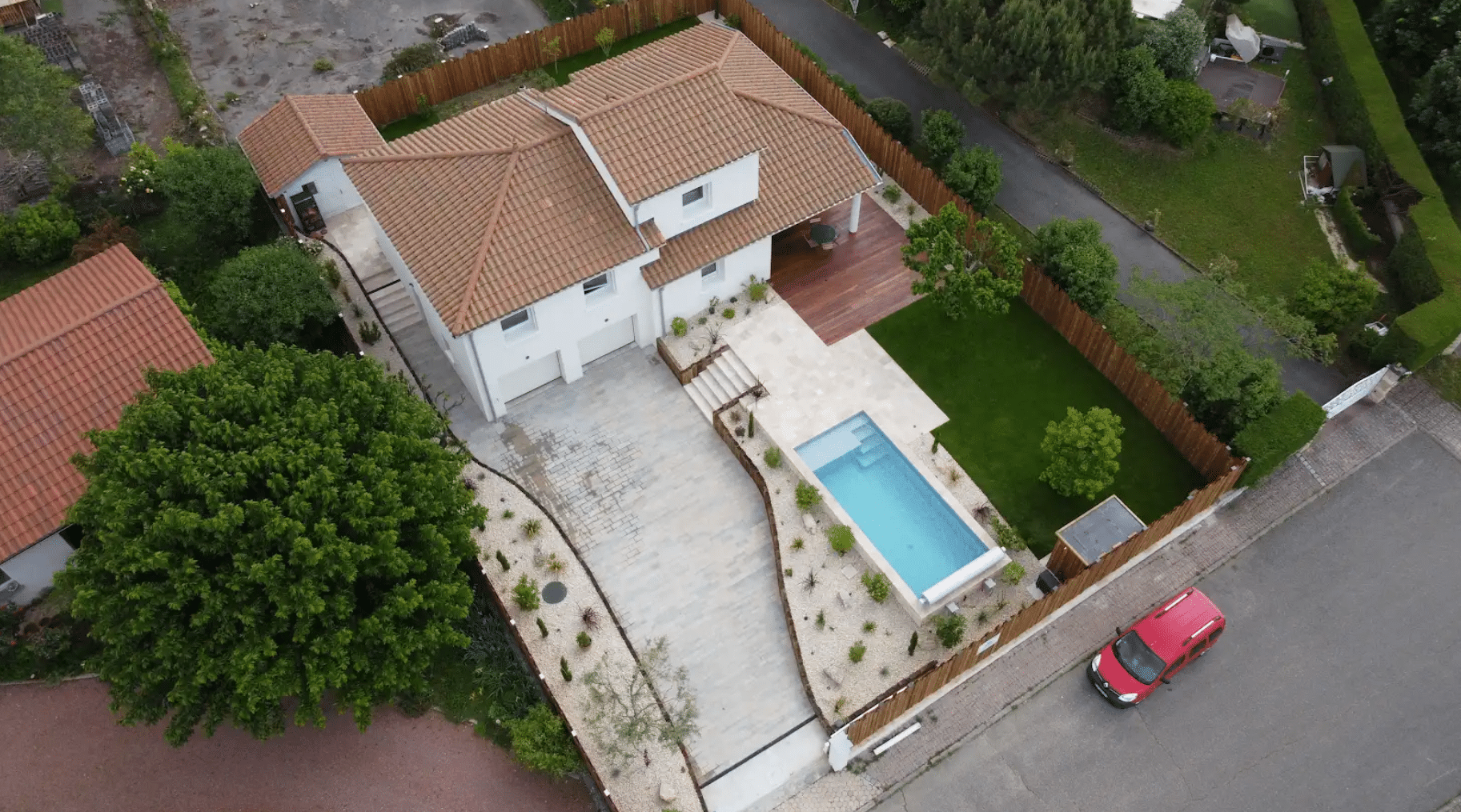 Maison moderne vue du ciel avec piscine rectangulaire, terrasse en bois, jardin paysager et voiture rouge devant.